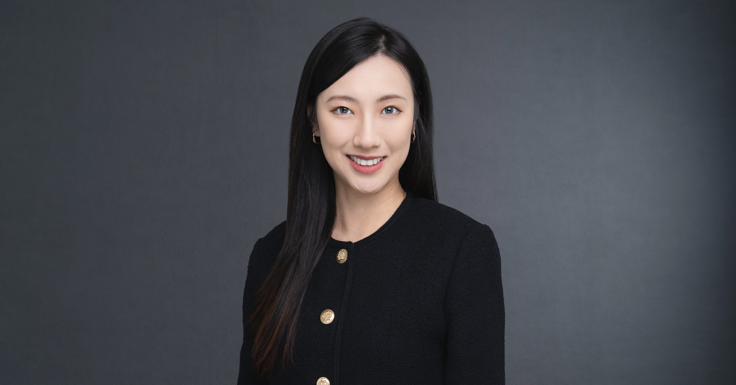 Portrait of an Asian woman with long black hair, wearing a black textured jacket with gold buttons, smiling at the camera.