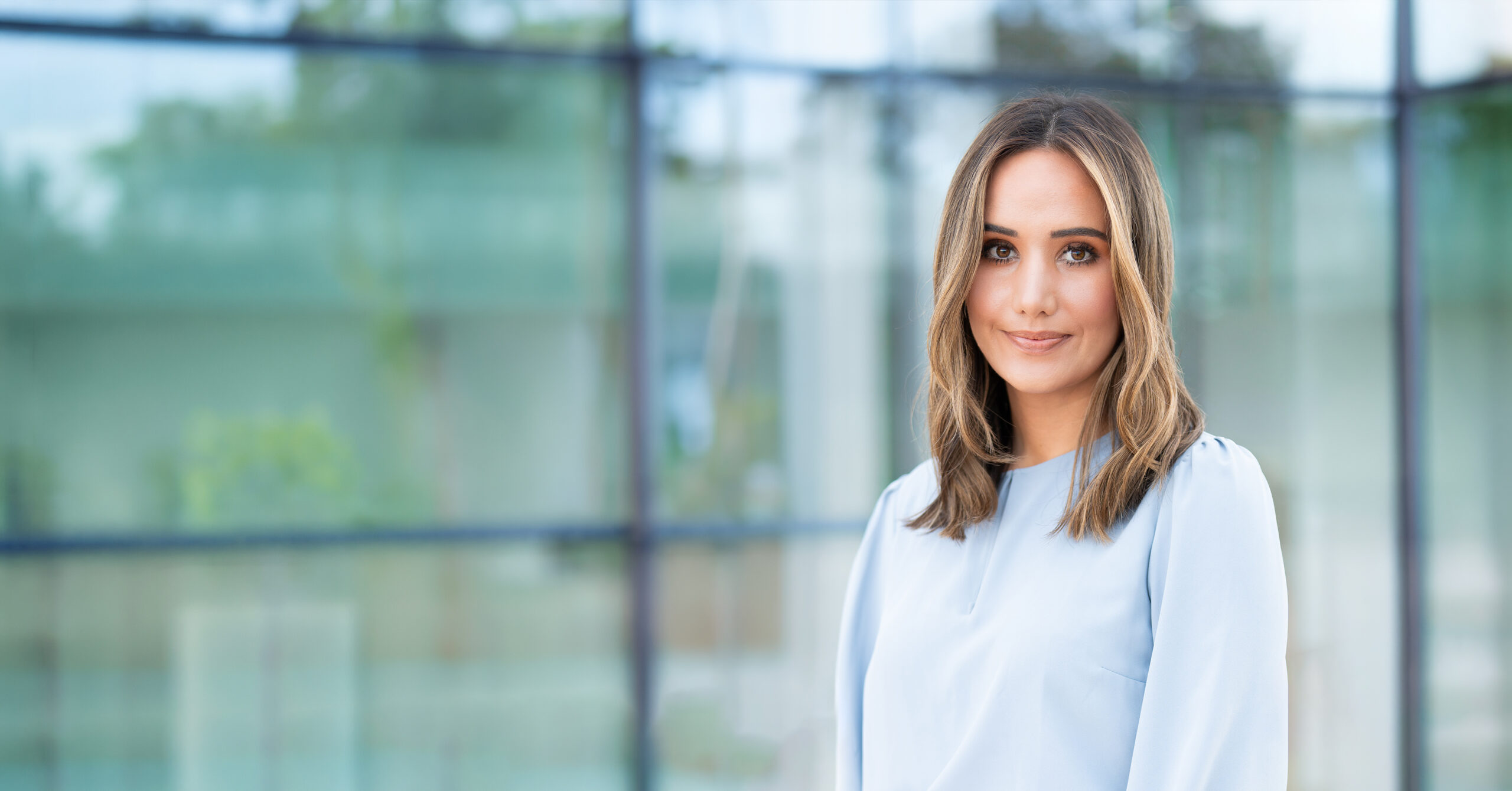 Professional woman standing outside a glass-front building, smiling gently at the camera, wearing a light blue blouse.