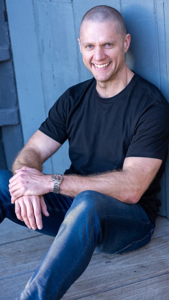 smiling bald young man in a black top and jeans sitting by a wooden fence