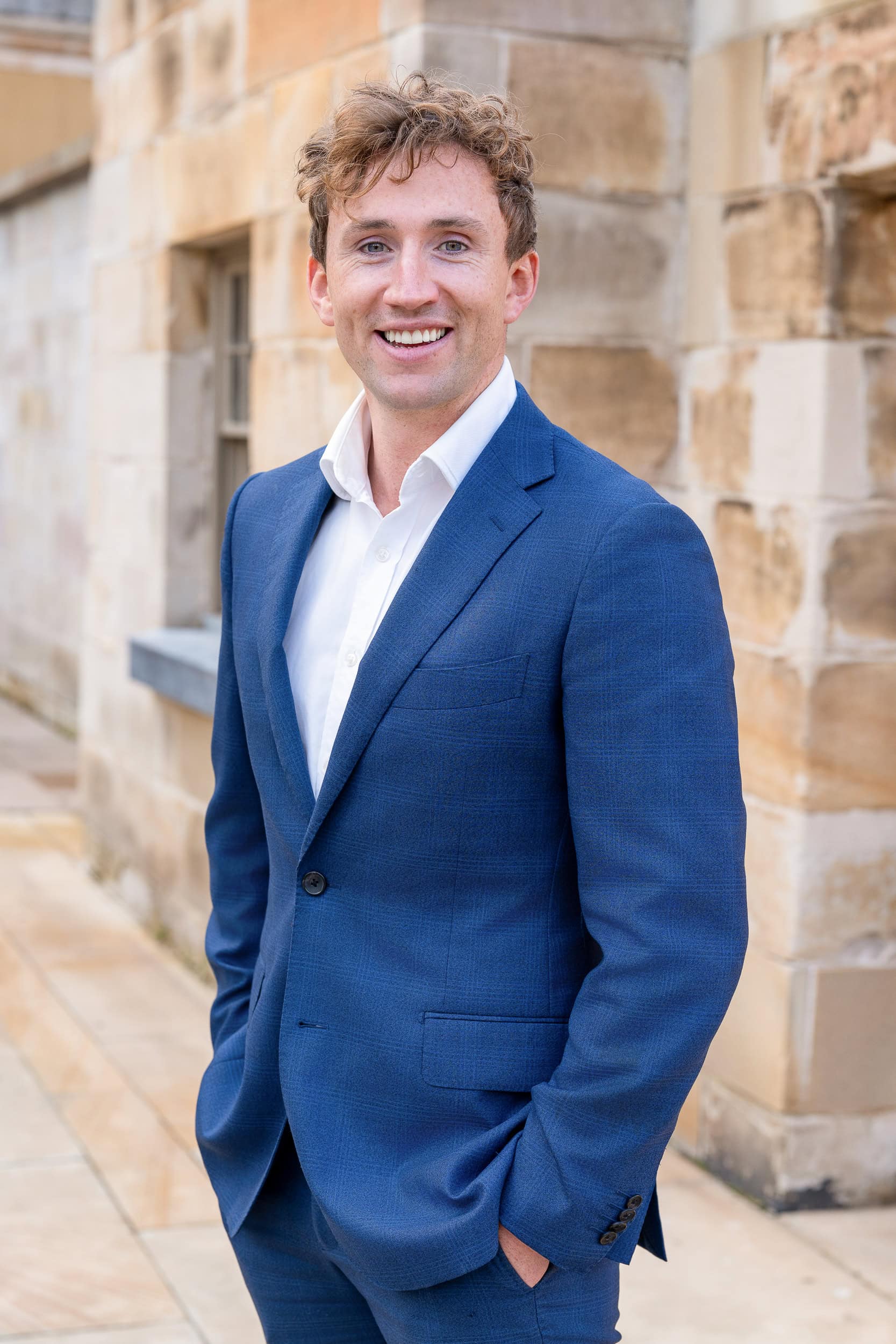 Portrait of a smiling man in a blue suit standing outdoors beside a stone building.
